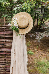 A beautiful straw hat with a bouquet of white flowers and a beige cape hangs on the fence. Nature and garden. Summer collection. V