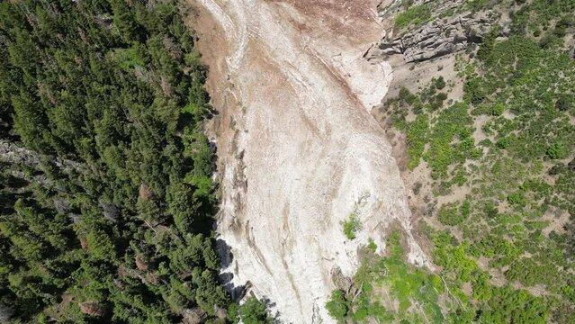 Aerial-Top down descending view over dirty debris strewn avalanche melting in spring-Mountains valley lines with pines