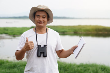 Handsome Asian man ecologist is surveying nature at the lake, holds paper notebook and binoculars....