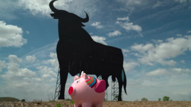 Hand of a man throwing coins into a unicorn piggy bank, with a typical Spanish landscape in the background, as a concept that in Spain or with a Spanish bank, the economy or savings grow.