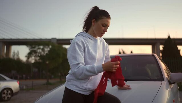 Tired female boxer wearing hoodie sitting on hood car and rests after evening outdoors boxing training. She has red boxing tape in her hands. Fit and healthy female resting after successful workout