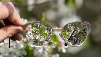 Myopia (shortsightedness) glasses in hand, looking on blooming spring trees garden in focus with blurry background. Nearsighted refractive lenses outdoors in nature. 