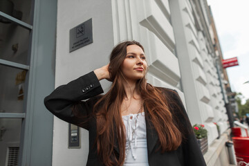 Happy fashion brunette woman model in stylish casual street outfit with white dress and black blazer walks in the city near a vintage building