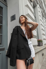 Fashionable young beautiful woman model in fashion clothes with white dress with black stylish blazer and handbag stands near a vintage white building outdoors