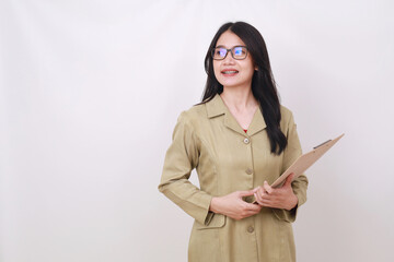 Pretty young Indonesian teacher standing while holding clipboard and looking sideways on empty space