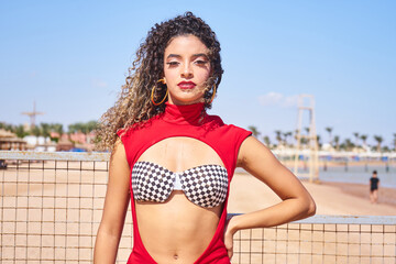 Portrait of sexy woman with slim figure in red suit and curly hair.Attractive Formula 1 beautiful model  Hostess posing on the beach.Bikini model