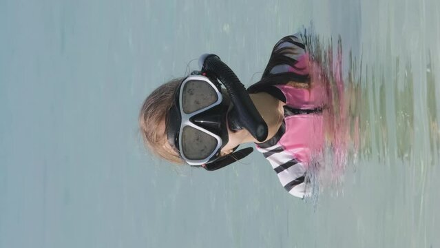 Vertical video. Adorable little girl in snorkeling mask waving hand at camera while swimming in the turquoise water of Indian ocean. Summer vacation and beach holiday concept