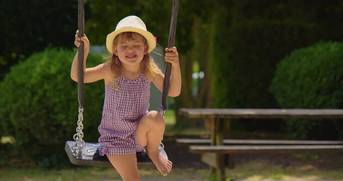 Little girl is swinging on a swing. Amusement park dream concept. Happy girl in hat swinging on swing in park at sunset. Portrait child plays with swing childhood dream. Baby swinging on swing in park