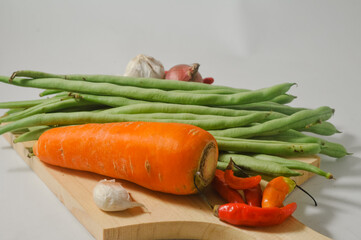 Some green beans, a carrot, some red chilies, some cloves of garlic, and an onion on a wooden cutting board isolated on a white background
