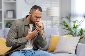 Young African American man sitting on the couch at home and coughing. Suffers from an attack of asthma, allergies. He holds his chest, covers his mouth with his hand - obrazy, fototapety, plakaty