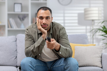 A young African American man is sitting on the couch at home and is bored. He lazily uses the TV...