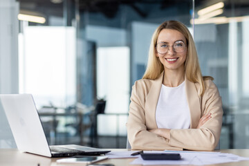 Portrait of a successful and self-confident business woman in a business suit sitting in the office at the desk and looking at the camera smiling with her arms crossed