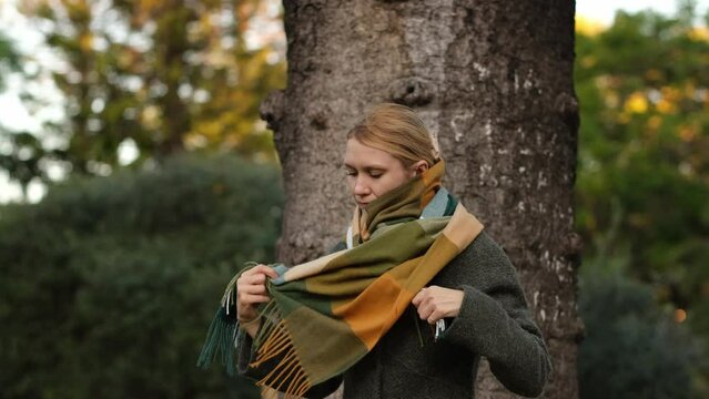 Blonde adult beautiful woman wearing coat tying a warm scarf by tree in park,half body.