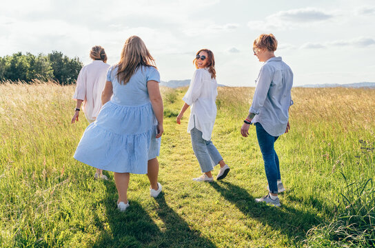 Four cheerful smiling women walking by the high green grass meadow hill, laughing and chatting with each other during a sunset walk. Woman's friendship, relations, and happiness concept image.