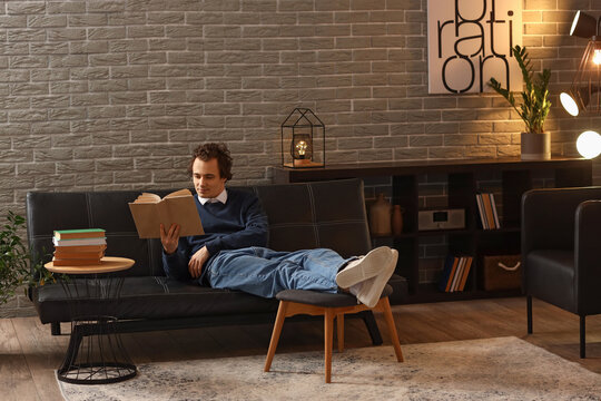 Young man reading book on sofa at home late in evening