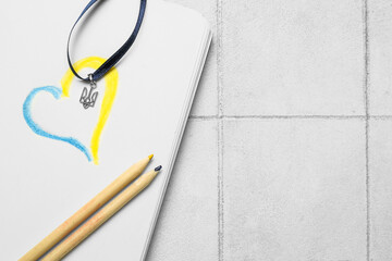Ribbon with Ukrainian coat of arms, notebook, drawn heart and pencils on white tile background