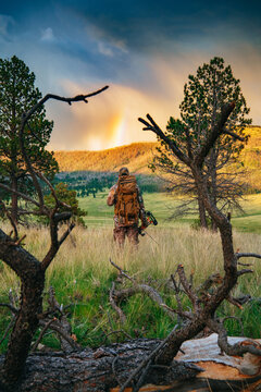 A hunter in camouflage and wearing a backpack watches a rainbow form under a storm while bowhunting in the Valles Caldera National Preserve, New Mexico