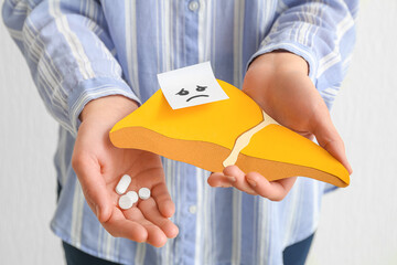 Woman holding yellow paper liver with gloomy face and pills on white background