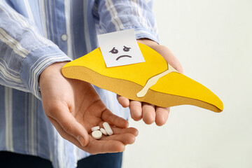 Woman holding yellow paper liver with gloomy face and pills on white background