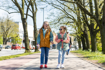 Two cute little children girlfriends enjoy fun talk walking in Hamburg city street park on sunny day outdoors. Happy small friends portrait wear sunglasses. Portrait of pair stylish kids outside