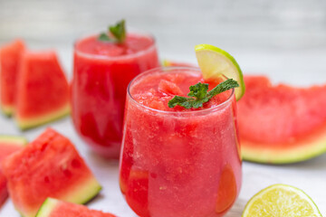 Watermelon smoothies with slices of fruit and leaf of mint and lime over white wooden background.