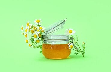 Glass jar of honey and fresh chamomile flowers on green background
