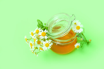 Glass jar of honey and fresh chamomile flowers on green background