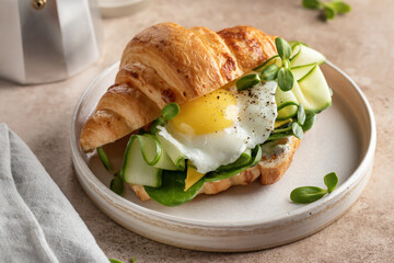 Delisious breakfast with croissant sandwich with fried egg, cheese, cucumber and micro greens and coffee over beige background close up