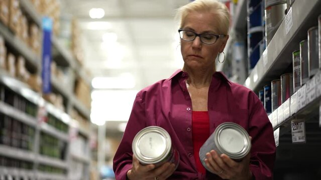 Attractive mature woman shopping for paint reads label and compares cans in a hardware store shopping for home renovation project.