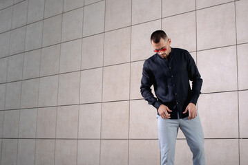 a stylish man in light pants and a black shirt on the background of an empty wall takes off something in sunglasses and looks down
