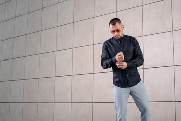 a stylish man in light pants and a black shirt on the background of an empty wall takes off in sunglasses and fastens the sleeve of his shirt