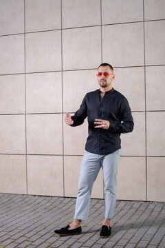 a stylish man in light pants and a black shirt on the background of an empty wall takes pictures in sunglasses shows a gesture with his hands