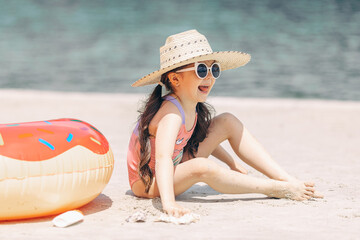 Side view of cute little girl sitting on the sand on a beach in a swimsuit and straw hat