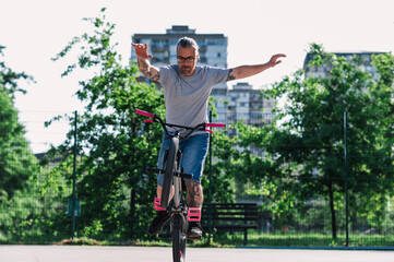 A trendy middle-aged tattooed man is practicing street-style bmx bike tricks in a skate park.