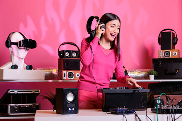 Asian musician with headphones playing electronic song at mixer console, standing at dj table having fun with fans at night in club. Artist performing techno music in studio over pink background