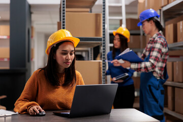 Postal service warehouse worker using parcel tracking system on laptop. Young asian storehouse employee wearing yellow hardhat managing product logistics and distribution on computer at desk