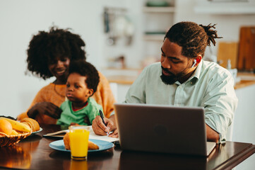 A focused multicultural man is having an online meeting on a laptop and writing tasks in the agenda while his wife is spending time with their son at the breakfast table.