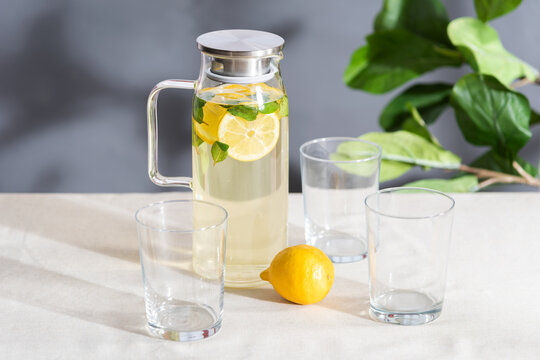 A pitcher of clear, refreshing water is surrounded by several glasses filled with slices of lemon and sprigs of mint