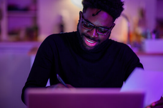 Portrait of an african american start-up businessman sitting at home late at night in a purple lighted room with his laptop and writing down in agenda. - Powered by Adobe