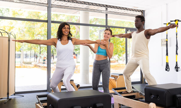 African-american man and latin woman doing exercises on reformers. Young caucasian woman pilates trainer correcting their moves.