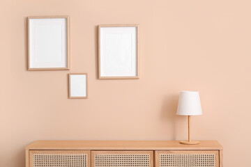 Modern chest of drawers with lamp and blank pictures hanging on beige wall in room