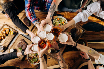 Fototapeta na wymiar Friends cheering beer glasses on wooden table covered with delicious food - Top view of people having dinner party at bar restaurant - Food and beverage lifestyle concept