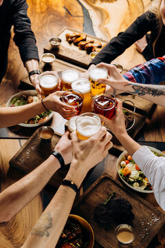 Friends cheering beer glasses on wooden table covered with delicious food - Top view of people having dinner party at bar restaurant - Food and beverage lifestyle concept