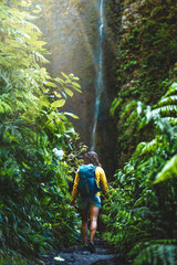 Backpacker woman walking along fern overgrown hiking trail torwards picturesque, overgrown waterfall in the Madeiran rainforest. Levada of Caldeirão Verde, Madeira Island, Portugal, Europe.