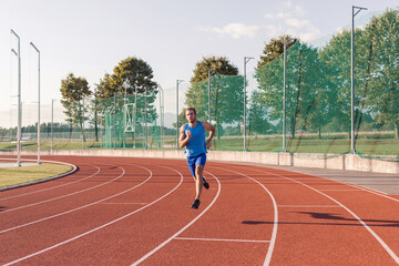 Caucasian male athlete in blue sportswear running alone along an athletic track on the stadium - obrazy, fototapety, plakaty
