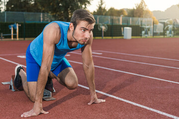 Young professional male athlete in blue sportswear taking a start position for a sprint run during outdoor training