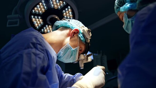 Busy neurosurgeon in device glasses at operation. Doctor using metal tool in his work. Female assisting.