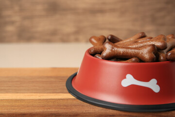 Red bowl with bone shaped dog cookies on wooden board, closeup. Space for text