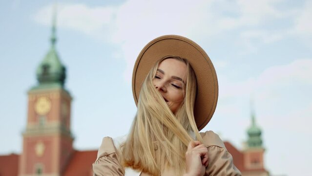 Portrait happy young woman wind blowing in long blond hair wearing fedora hat stands in front of building in historical part of city on sunny day. Cute female looking at cityscape and smiles to camera