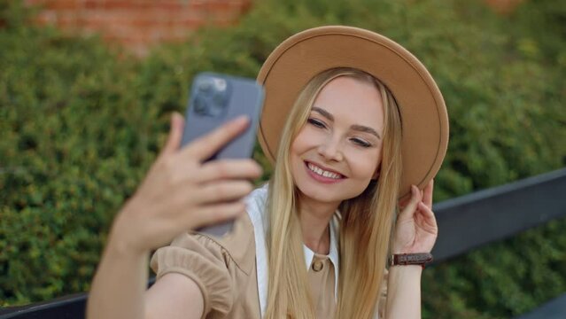 Young woman wearing brown sun hat sitting on park bench taking selfie with smartphone in hand. She is smiling looking at cell phone screen. Summer season, red brick wall and green bushes on background